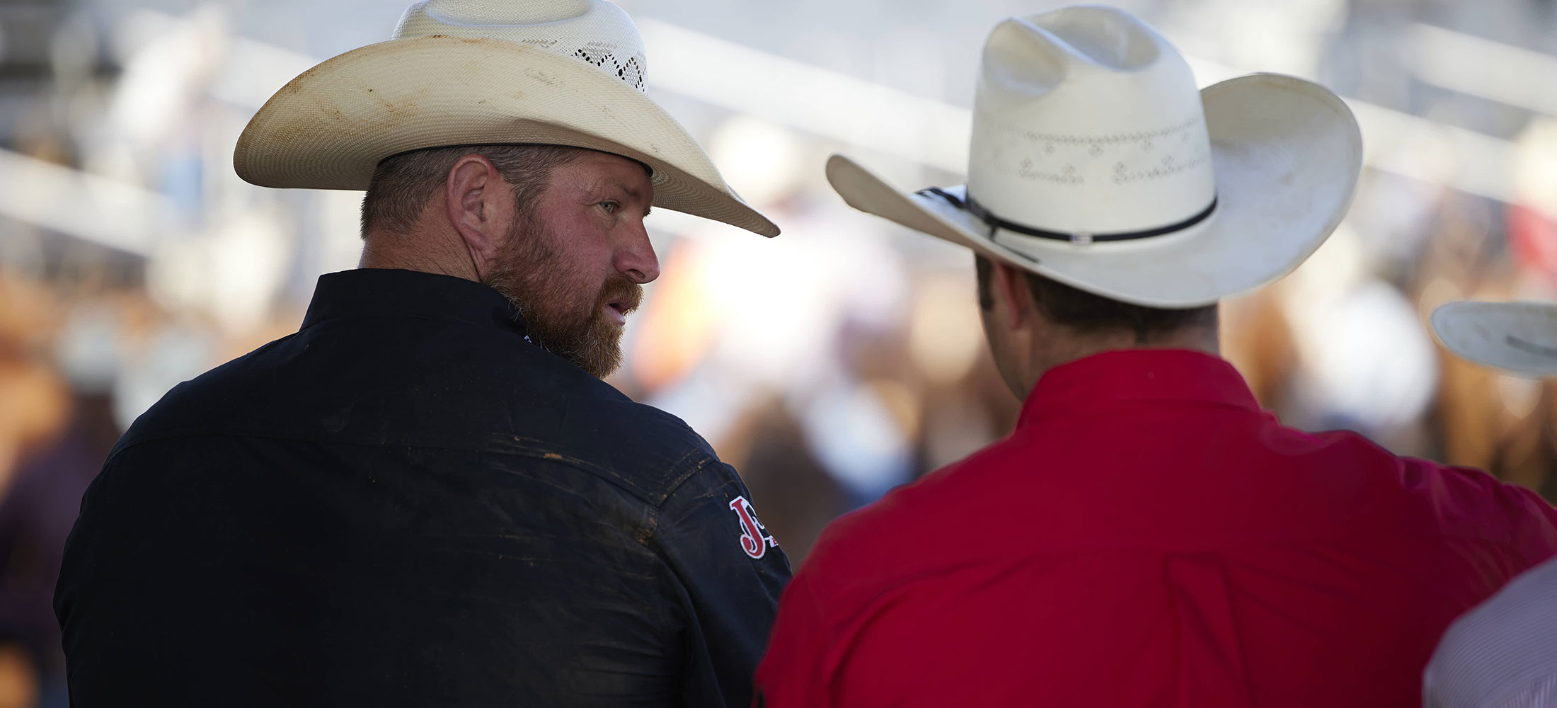 A cowboy wearing a black shirt looking at a cowboy wearing a red shirt at a rodeo.